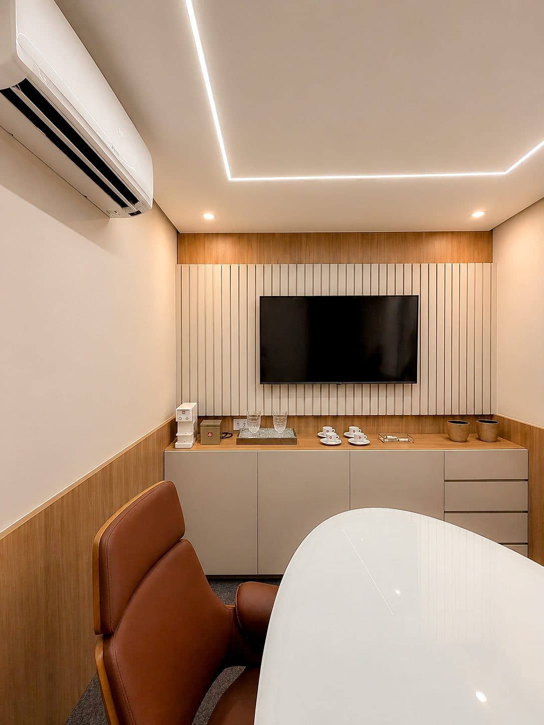 Sala de reunião pequena do MYJOB Executive vista da cabeceira: mesa oval branca, cadeiras de couro marrom, TV no painel ripado e aparador com xícaras
