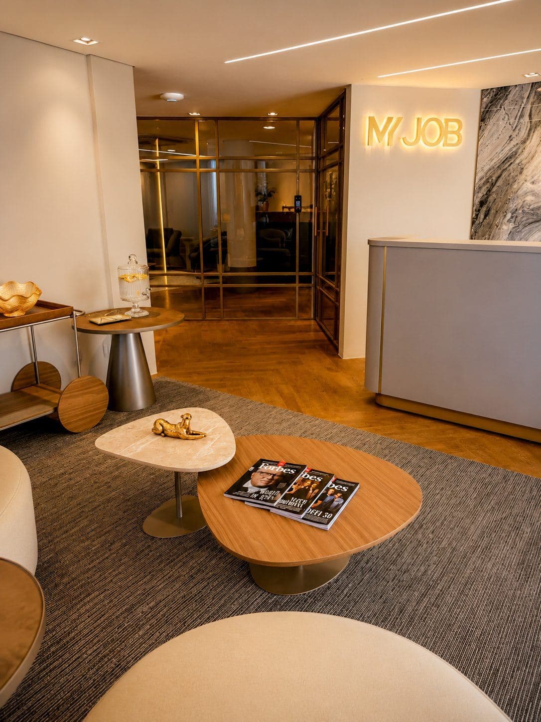 MYJOB Afonso Pena: recepção com o letreiro MYJOB iluminado, balcão cinza com filete dourado, painel de mármore, divisória de vidro e mesas de centro com revistas em primeiro plano